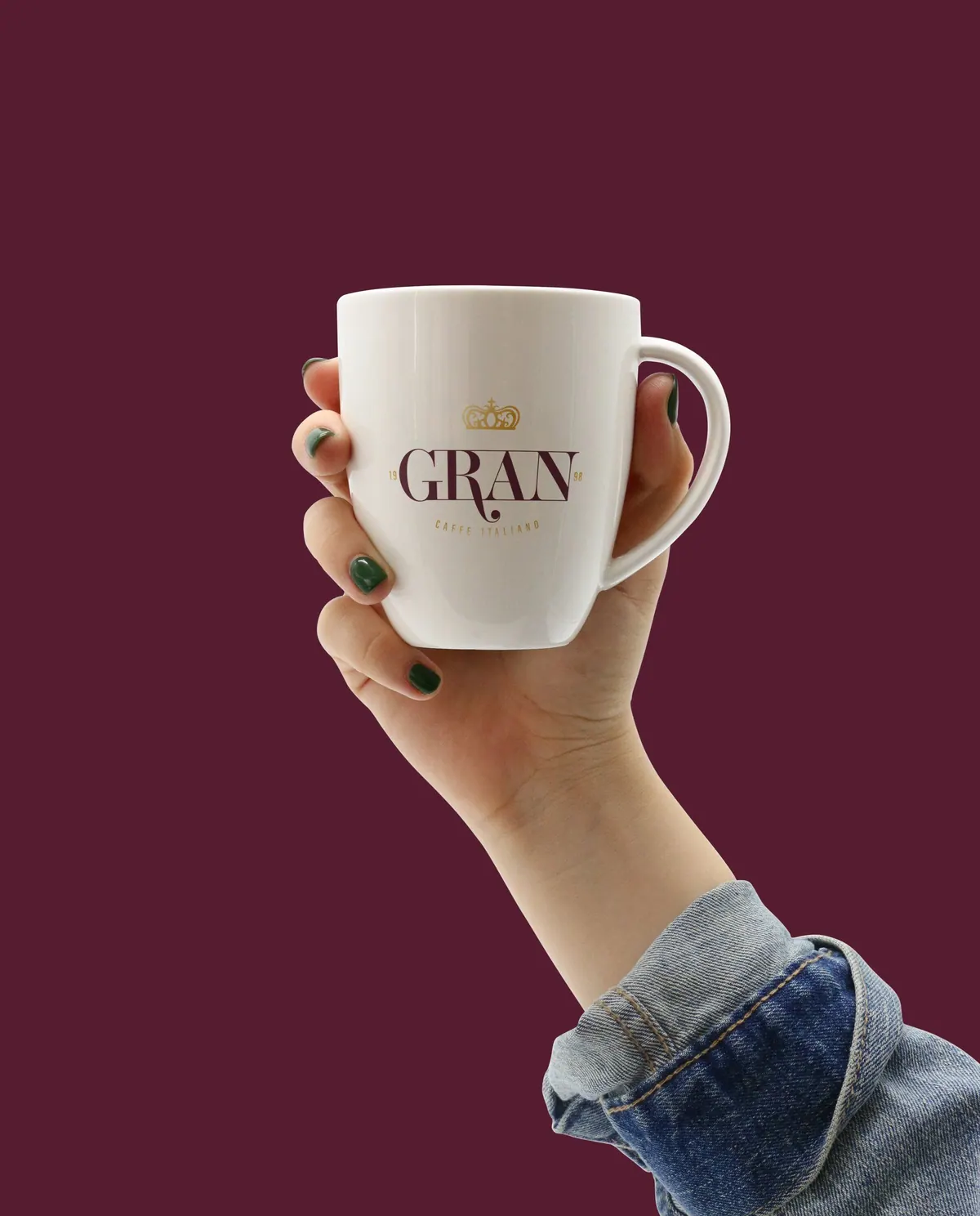 Χέρι κρατά λευκή κούπα με το λογότυπο GRAN Caffè Italiano: χρυσό στέμμα, GRAN σε μπορντό και 19 98, σε μπορντό φόντο